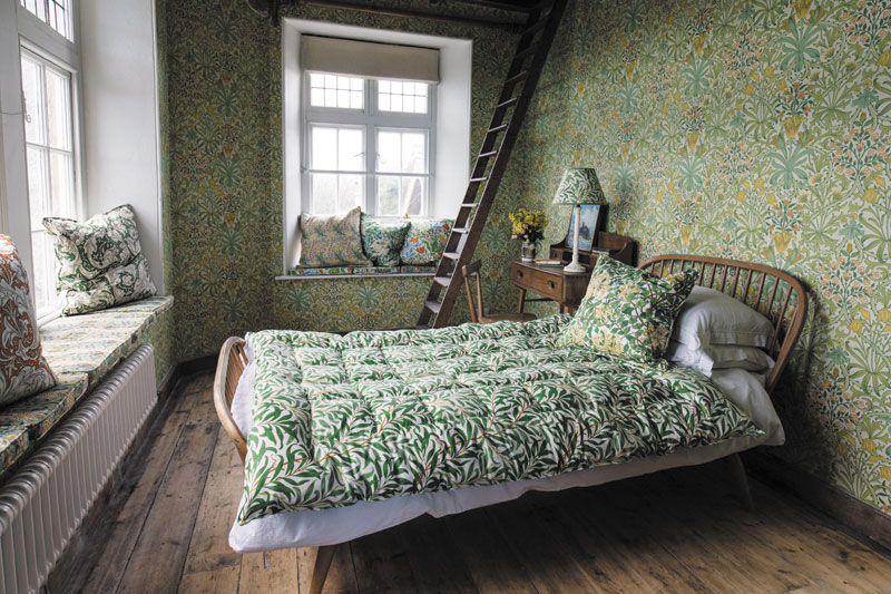 dormitorio con colcha, cojines, papel pintado para pared y lámpara con motivos vegetales