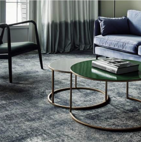 alfombra personalizada en tono gris en habitación de hotel junto a sofá tapizado azul  y cortinas degradadas