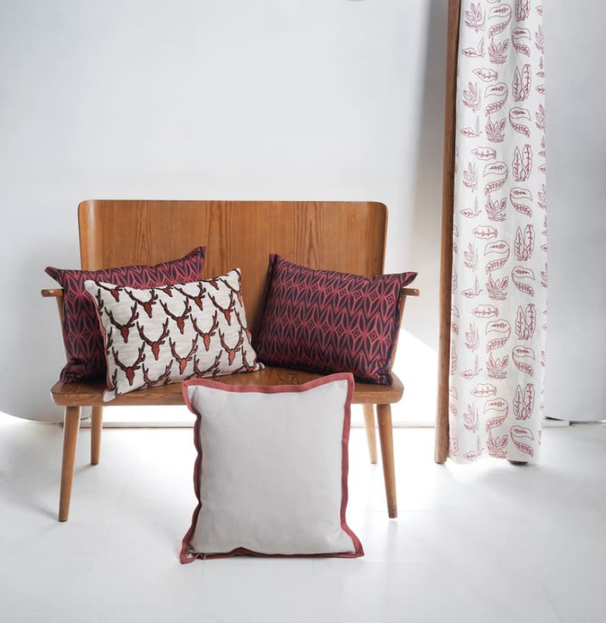 sillón de madera con cojines en diseños geométricos, estampado de ciervos y liso en tonos caldero y beige junto a cortina con estampado floral en tono caldero