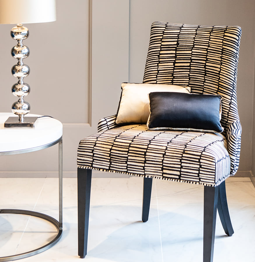 detalle de habitación sillon tapizado con tejido geométrico en tono beige y negro, cojines lisos a juego y mesa y lámpara de pie en tonos plateados y blancos