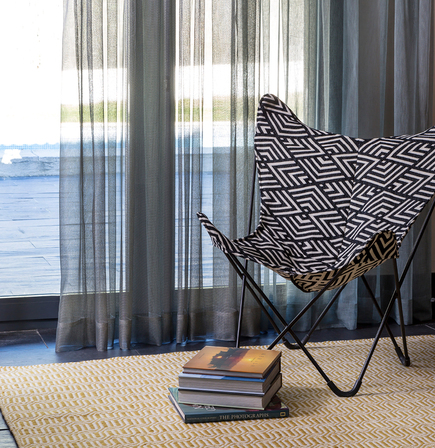 habitación con cortinas grises y sillón con tela geometrica negra y blanca, debajo alfombra geométrica en tonos amarillo y beige